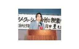 演壇に立ってマイクの前で話をしているスーツ姿の指田豊博士の写真