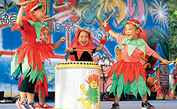 4人の子ども達が赤や緑を基調とした衣装を身にまとい、舞台上の中央に置かれた太鼓をたたいている様子の写真