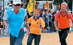 年配の男性と女性がハチマキやゼッケンを付けて、運動場でリレー競争をしている写真