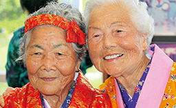 2人の年配の女性が鮮やかな赤とオレンジ色の着物を着て、にこやかな表情をしている写真