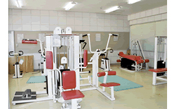 部屋の壁際や中央に、白を基調とし座面などが赤いトレーニング機器が、スペースを開けて置かれている大宜味小学校クラブハウスのトレーニング室の写真
