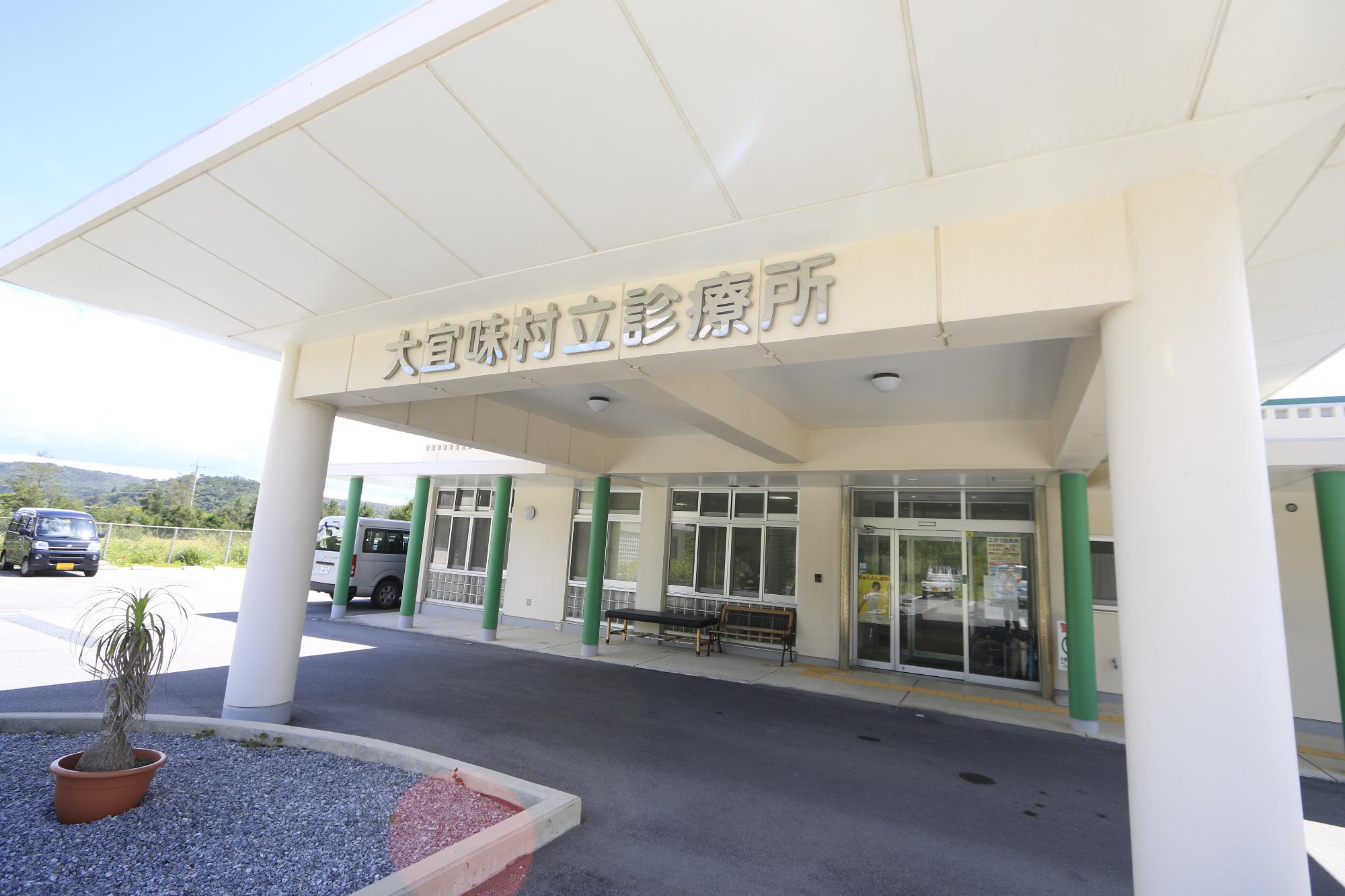 白を基調とした建物で柱が緑色になっており、入口にベンチが設置されている診療所入口の写真